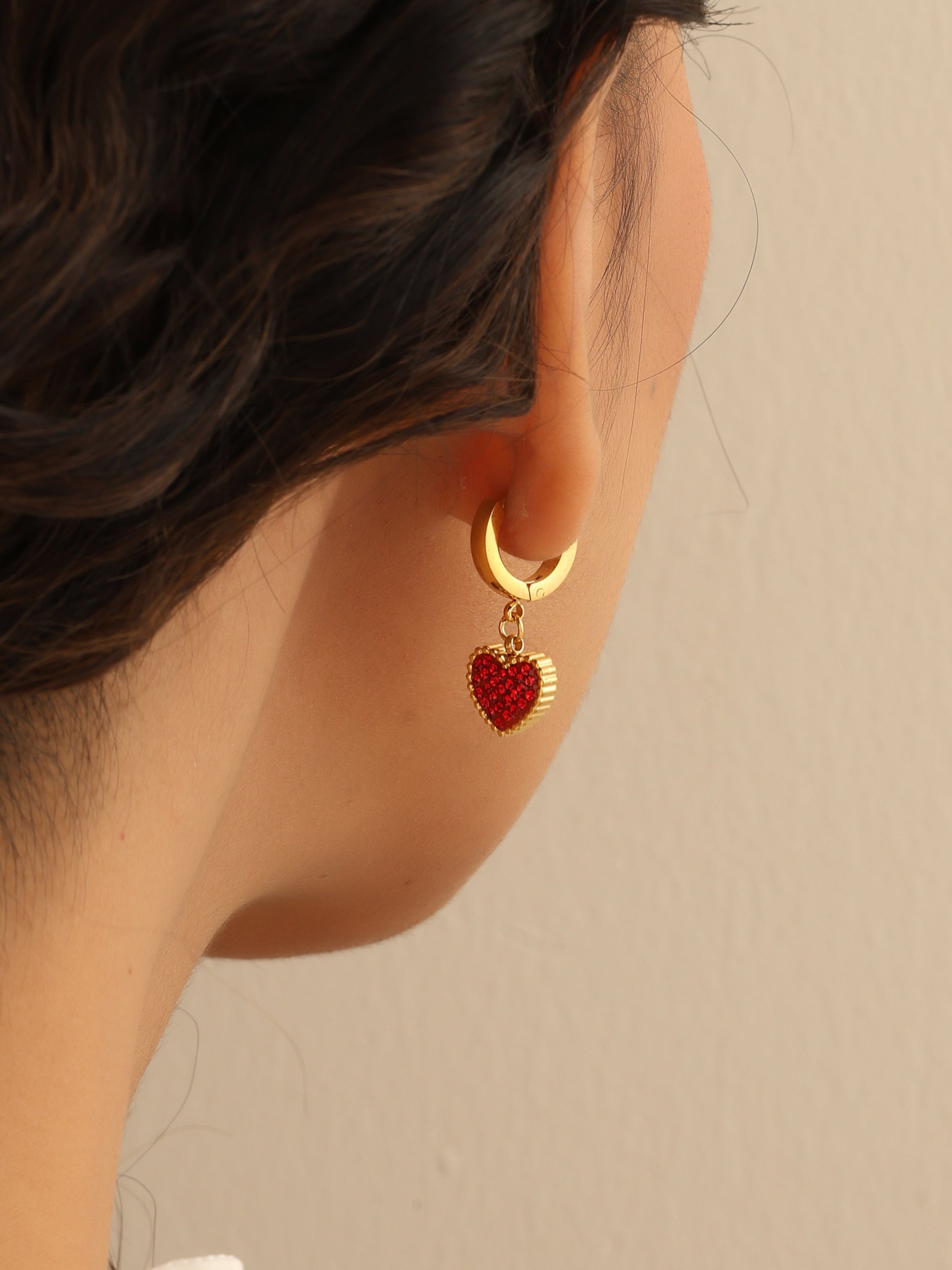 Close-up of a person wearing gold hoop earrings with red heart charms against a beige background