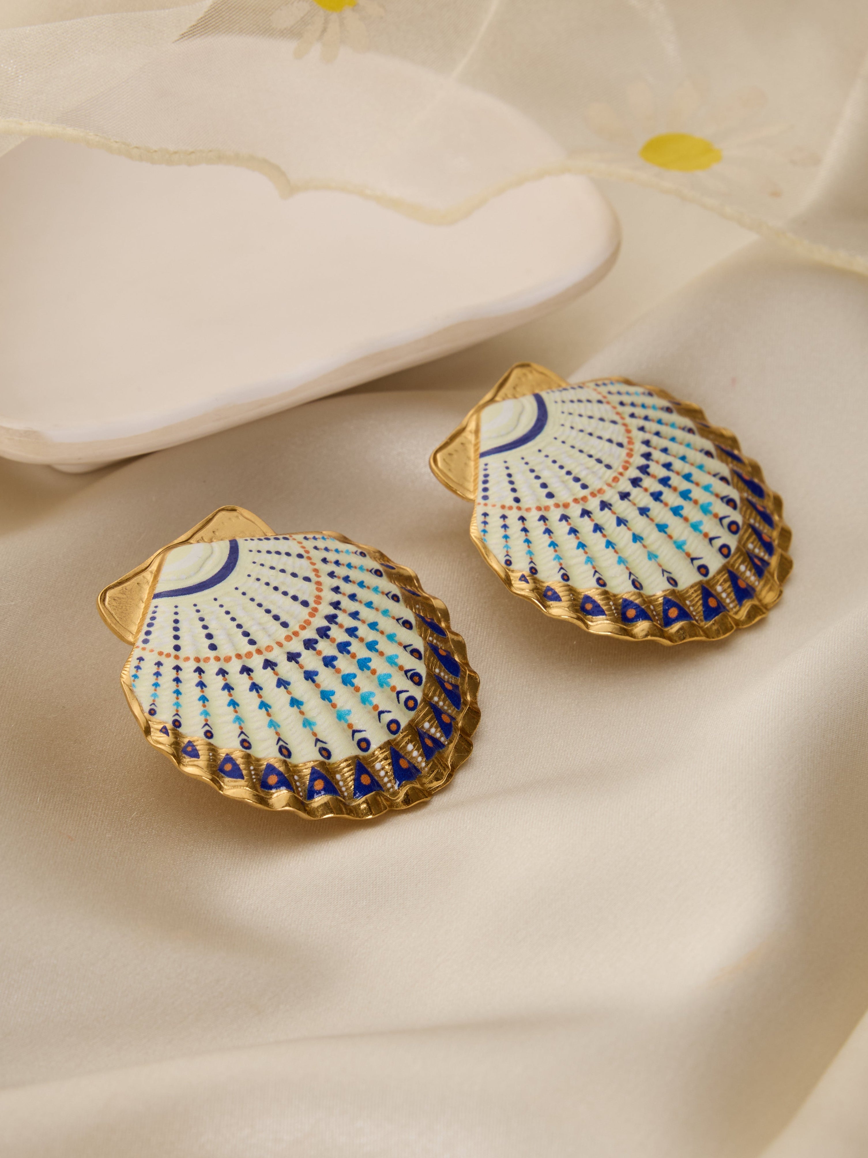 Shell-shaped earrings with blue and gold patterns on a beige fabric background
