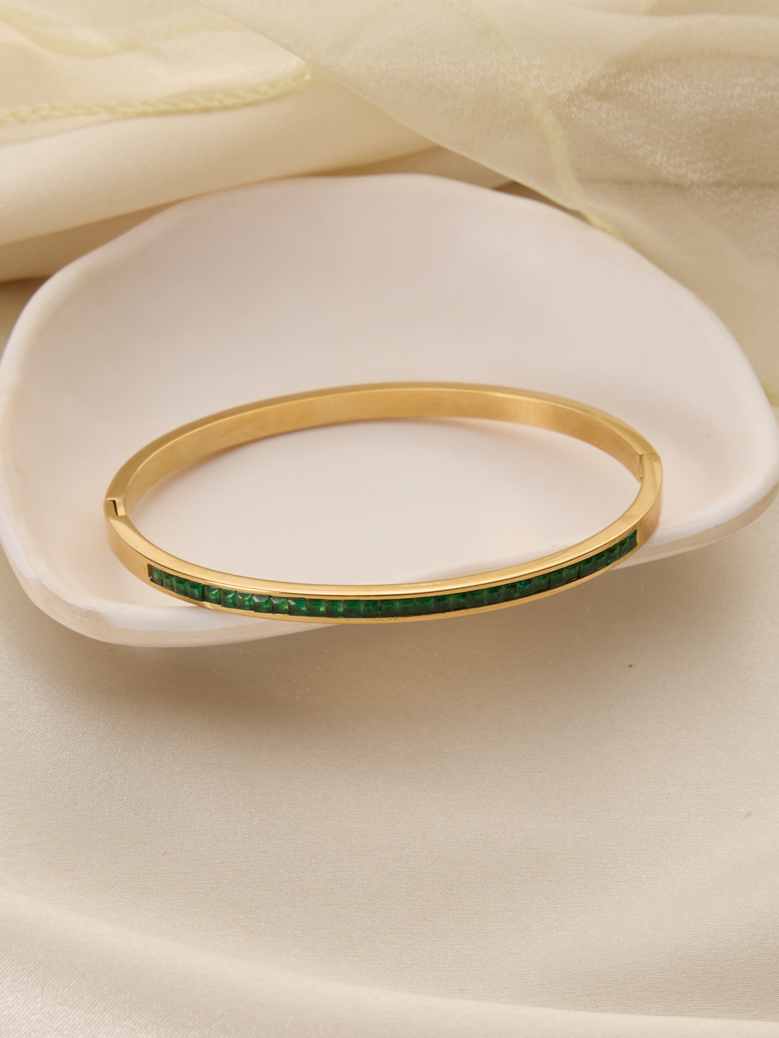 Gold bracelet with green stones on a white dish with beige fabric background