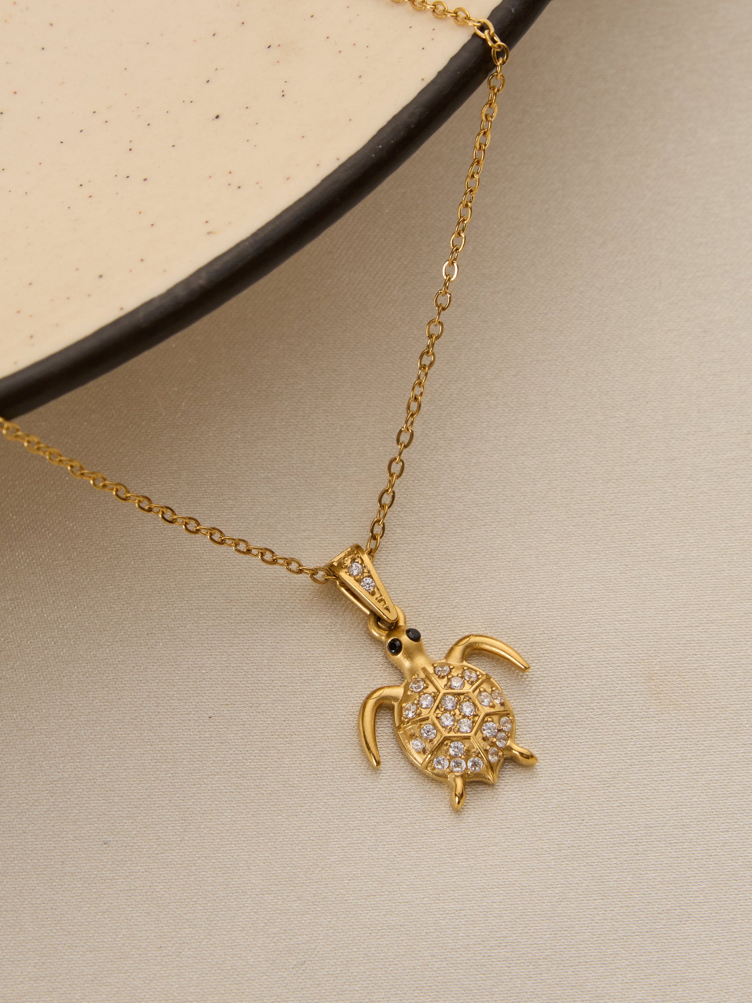 A gold chain necklace with a sea turtle pendant paved with small clear stones, displayed on a textured beige surface with a black-rimmed plate.