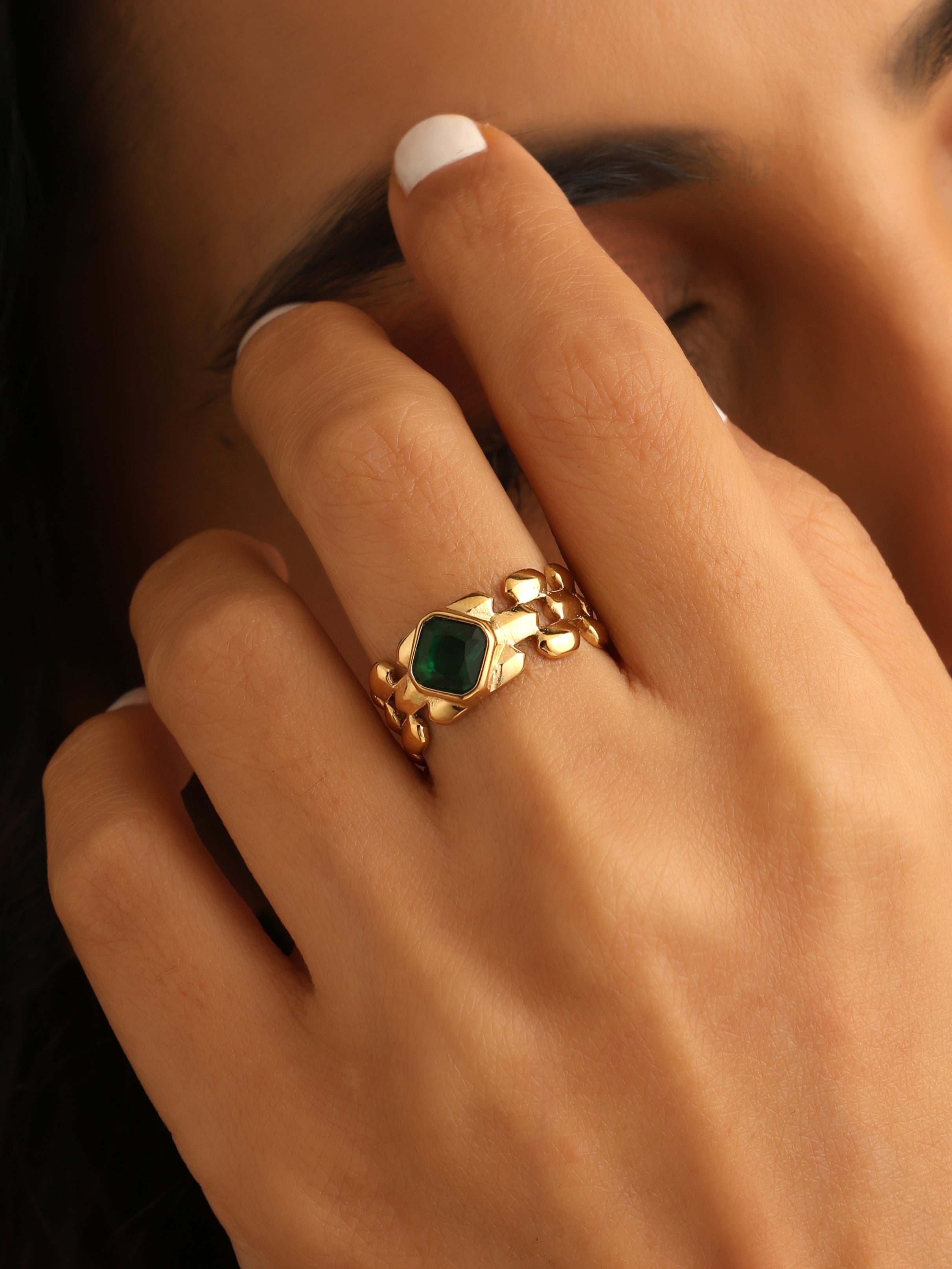 Close-up of a hand wearing a gold ring with a green gemstone, touching the face.