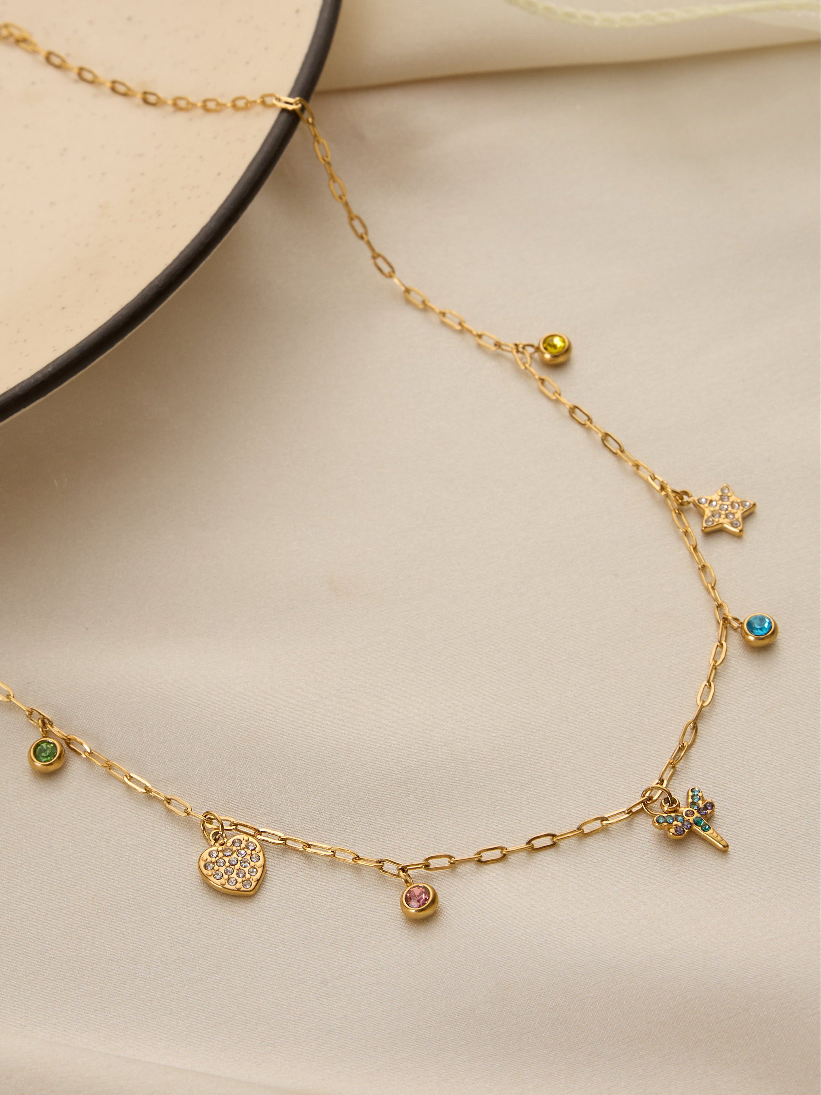 A delicate gold chain necklace with multiple small charms, including a heart, a flower, a star, and tiny bead accents in green, yellow, and blue, displayed on a soft beige fabric background.