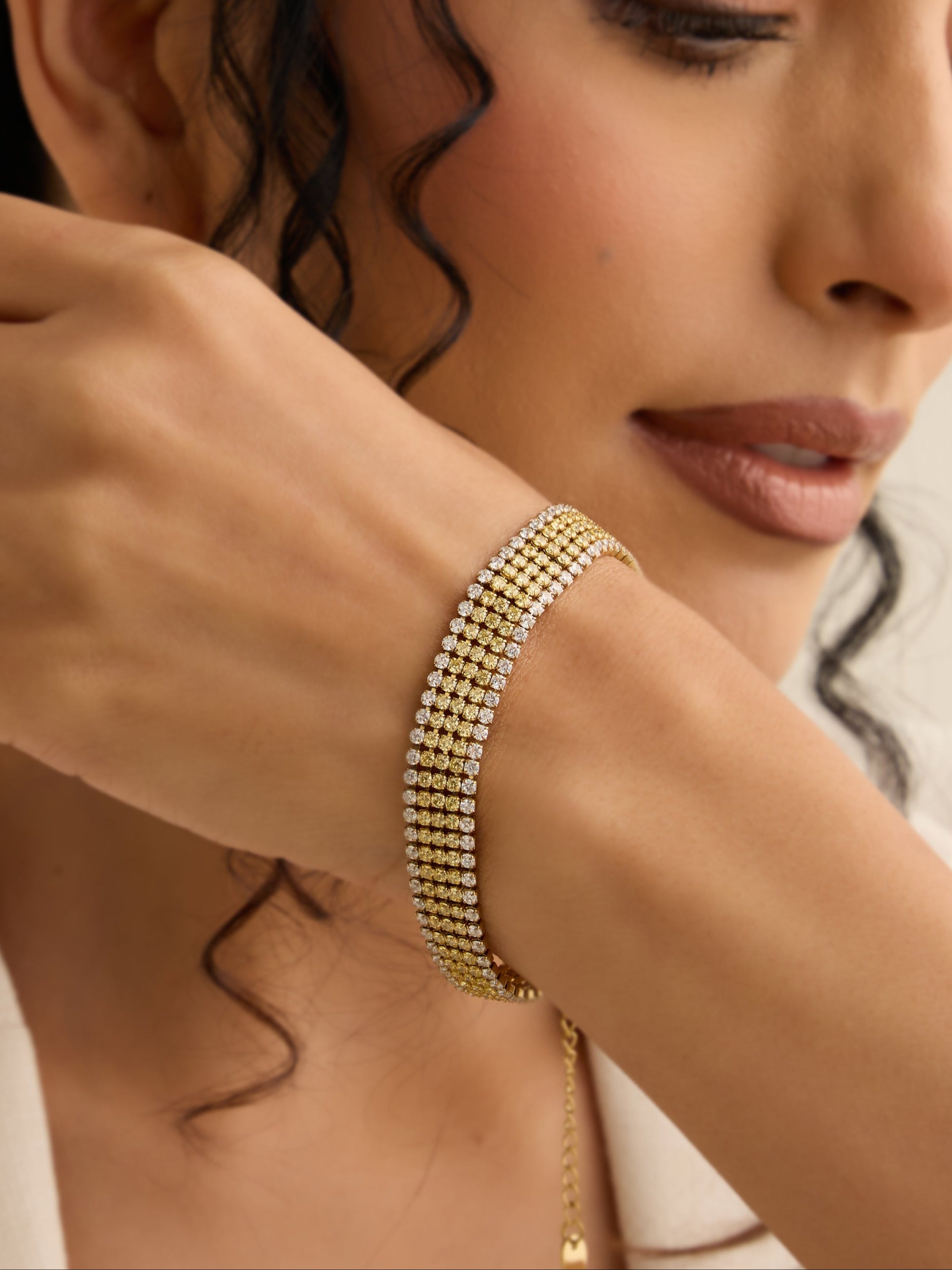 Woman wearing gold jewelry, including a bracelet and necklace, against a neutral background.